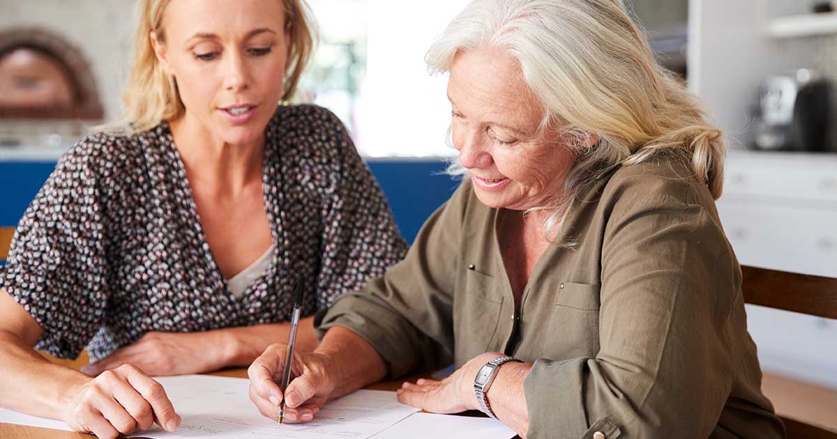 Female Friend Helping Senior Woman To Complete Last Will And Testament At Home