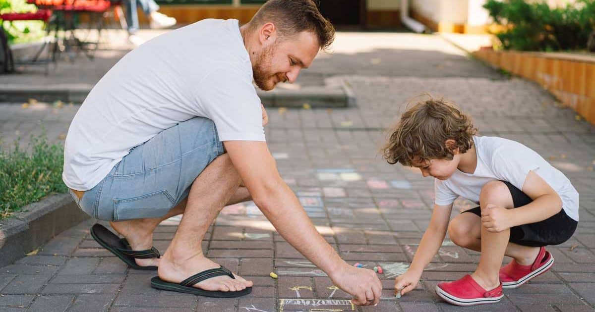 A little preschool boy with father draws with colorful chalks on the ground