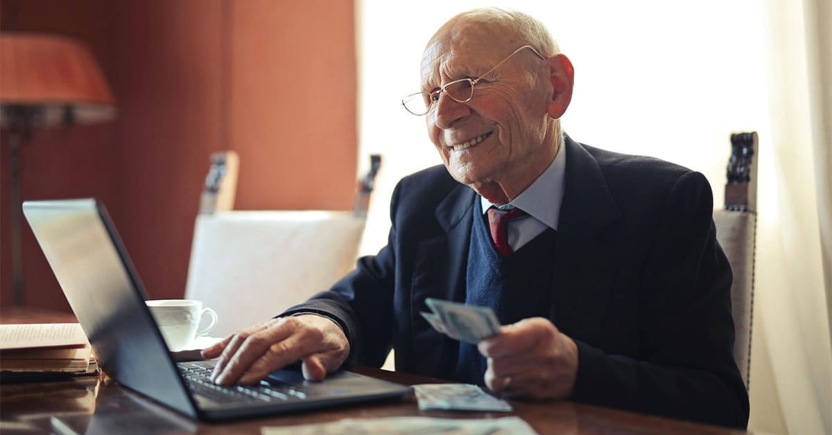 elderly man using a laptop and holding dollar bills