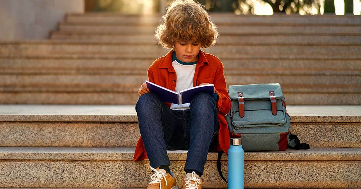 Back to school. A cute child with a backpack sits on the stairs near the school and reads a textbook. Elementary school student studying homework after class.